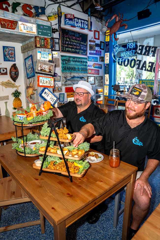 Two staff members at a wooden table inside Big Water Fish Market with a tiered seafood tower, beneath walls covered in out-of-state license plates and a neon Fish Market sign